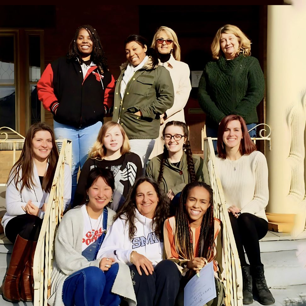 A group of smiling women poses together on steps, inviting others to join their supportive doula community.